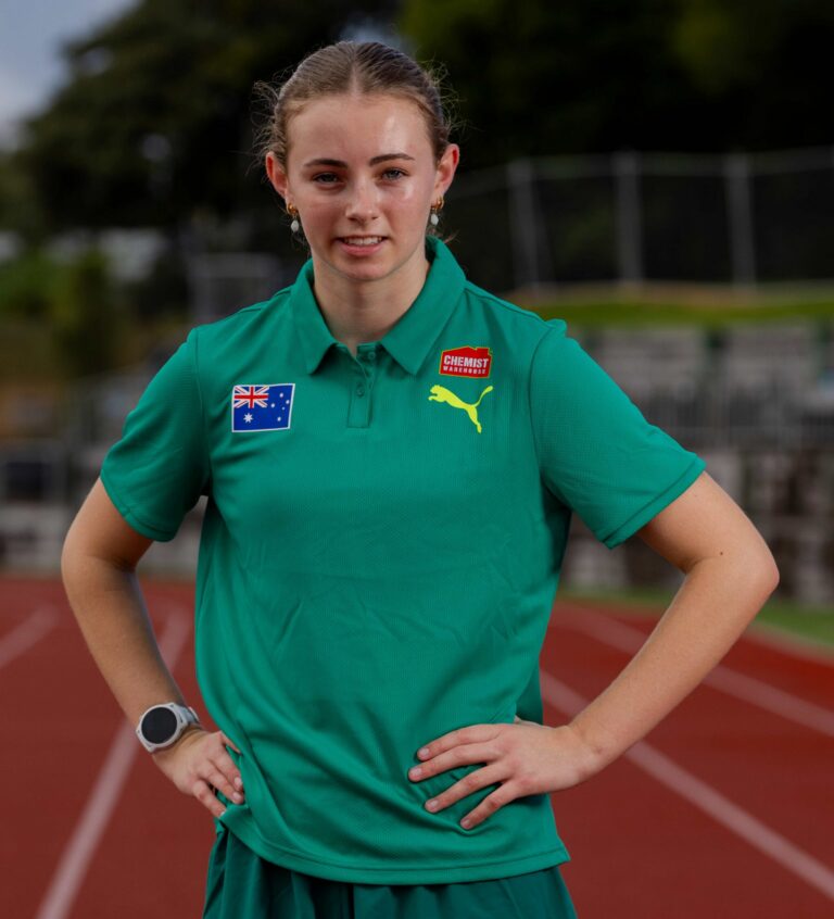 Young woman standing on a running track with hands on hips, wearing a green polo shirt with Australian flag and Puma logos visible.