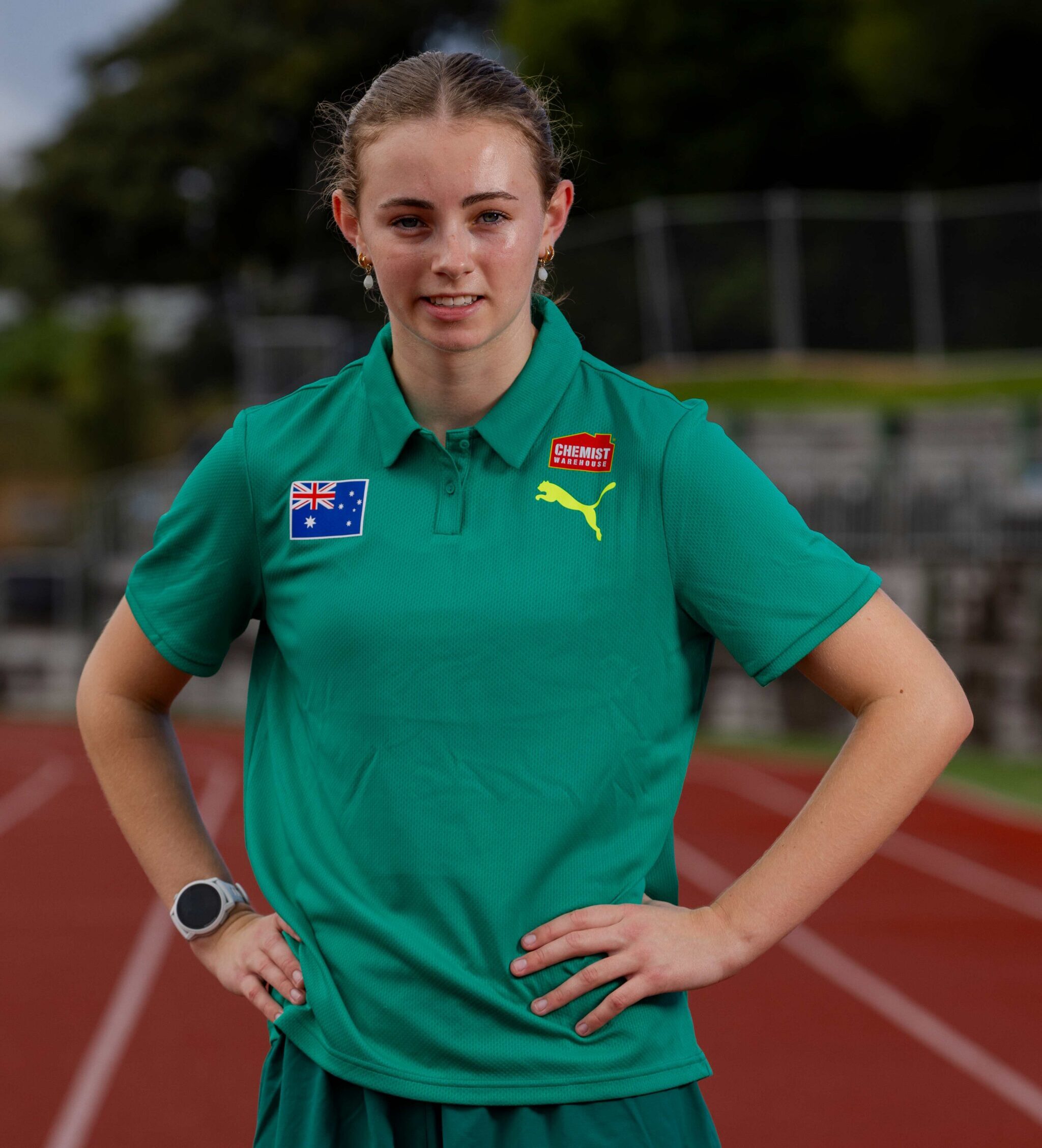 Young woman standing on a running track with hands on hips, wearing a green polo shirt with Australian flag and Puma logos visible.