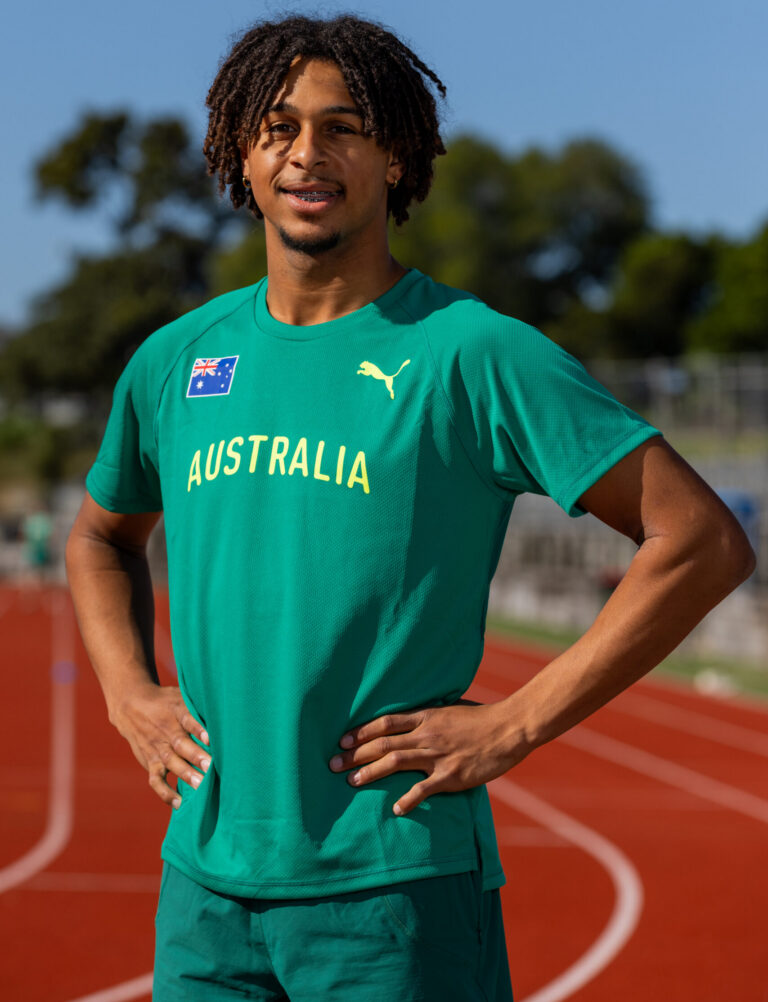 Young man on an outdoor running track wearing a teal Australia jersey, hands on hips, blue sky and trees in the background.