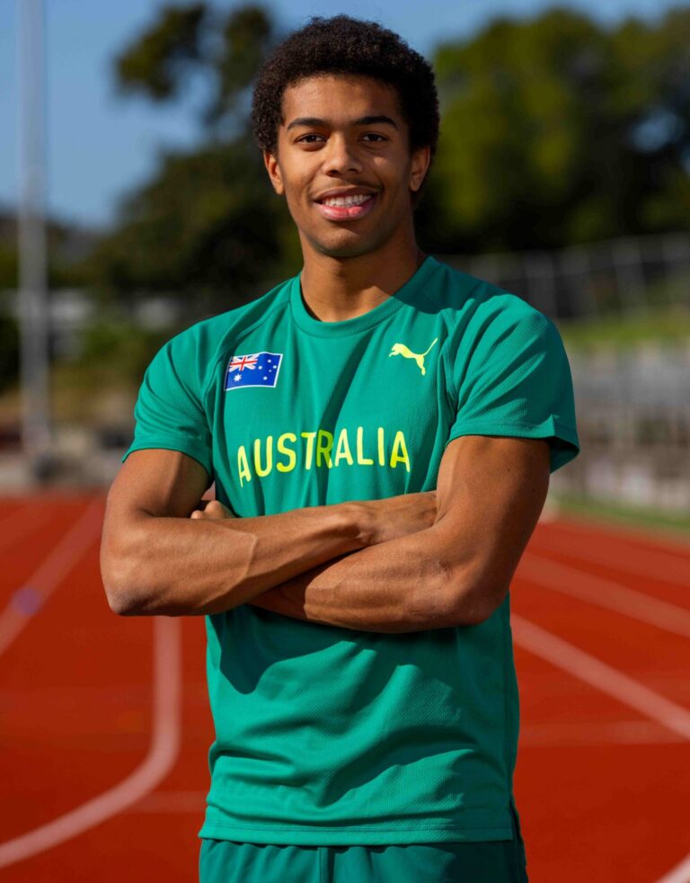 Smiling young man in a green Australia jersey stands on an outdoor running track, arms crossed.