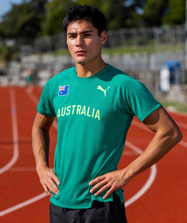 Young man in a teal Australia training shirt standing on a running track, looking at the camera.