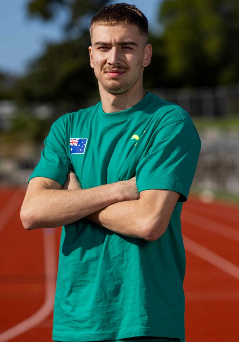 Young man with short dark hair wearing a teal athletic shirt stands with arms crossed on a sunny outdoor running track.
