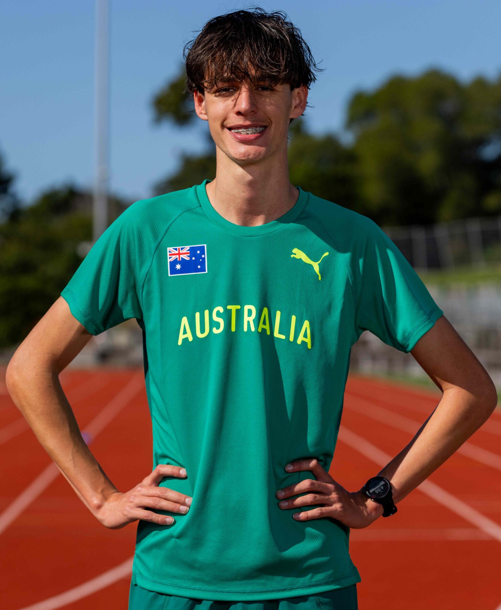 Young athlete standing on a track in a green Australia shirt, hands on hips and smiling at the camera.