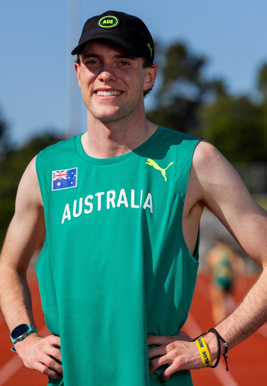 Smiling male athlete standing on a track, wearing a green Australia jersey with the Australian flag and 'AUSTRALIA' text, and a black cap with AUS logo.