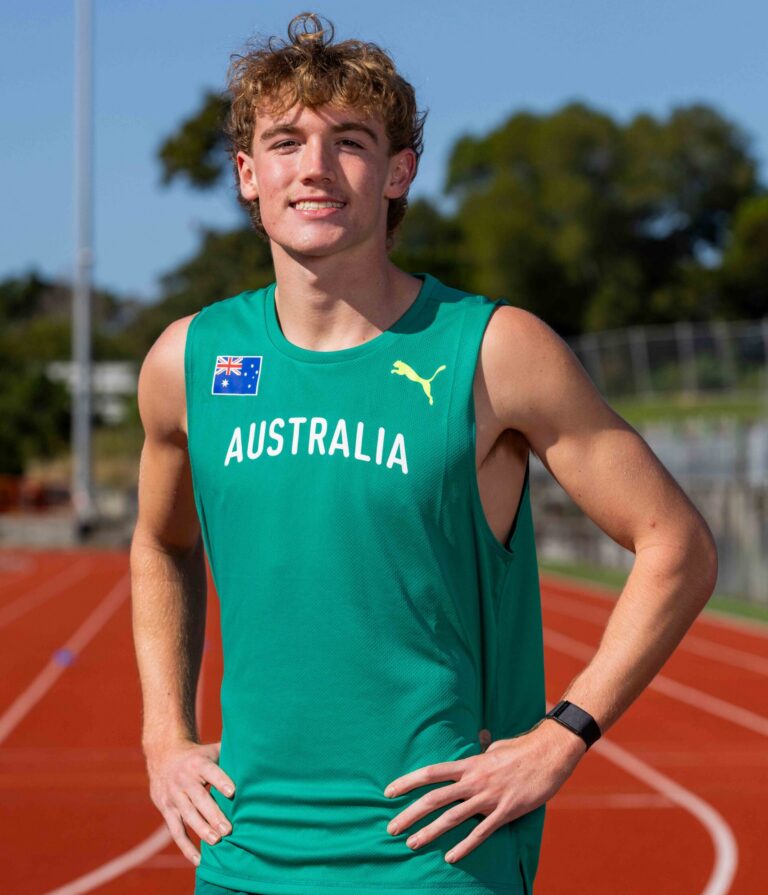 Young male athlete on a running track wearing a teal Australia jersey, hands on hips, smiling at the camera.