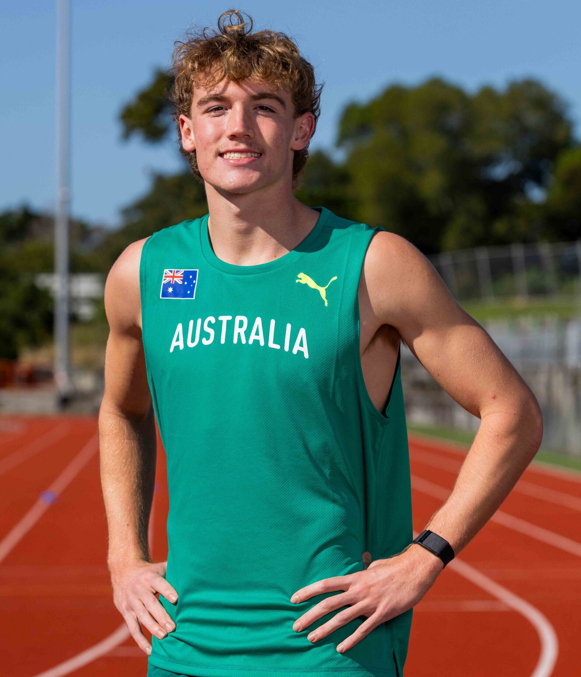 Young male athlete on a running track wearing a teal Australia jersey, hands on hips, smiling at the camera.