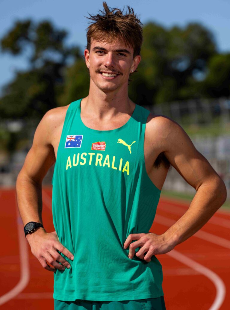 Male runner in a teal Australia singlet stands on a track, smiling at the camera with hands on hips.