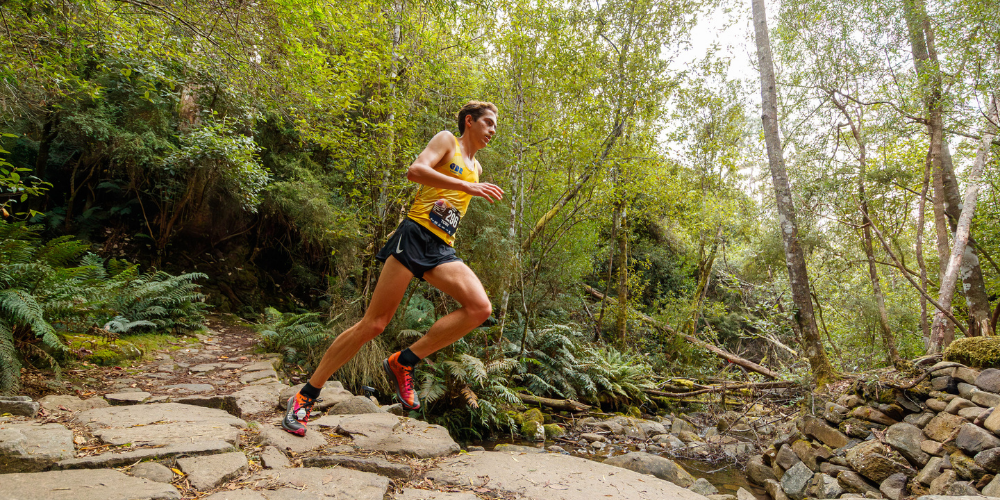 Male trail runner in a yellow sleeveless shirt and black shorts mid-stride on a rocky forest path, with a race bib on his chest.