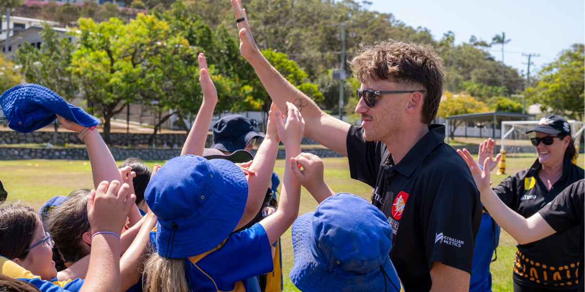 Coach in sunglasses high-fiving a group of children in blue hats on a sunny sports field, celebrating a drill.