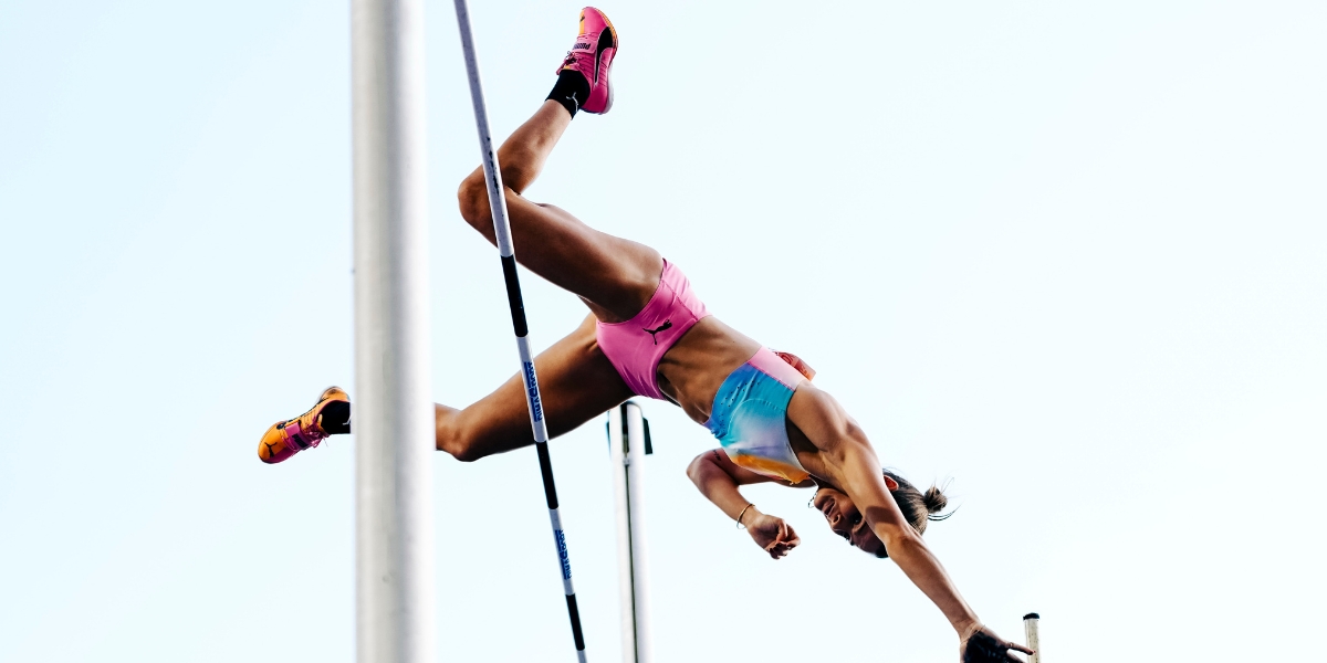 Female pole vaulter in colorful gear mid-air during a vault, holding the pole as she clears the bar on a sunny day, sky backdrop