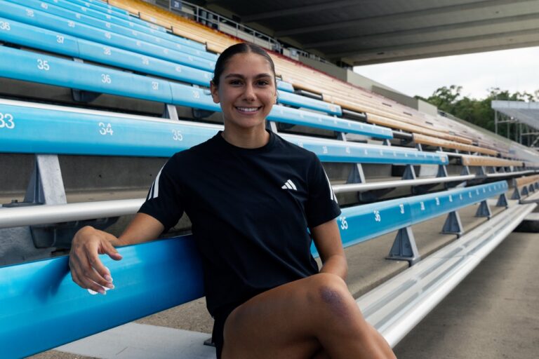 Smiling woman in a black athletic shirt sits on blue stadium bleachers, posing for a photo outdoors.