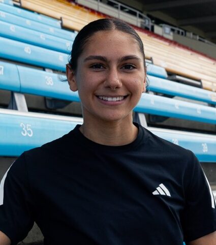 Smiling woman in a black athletic shirt sits on blue stadium bleachers, posing for a photo outdoors.