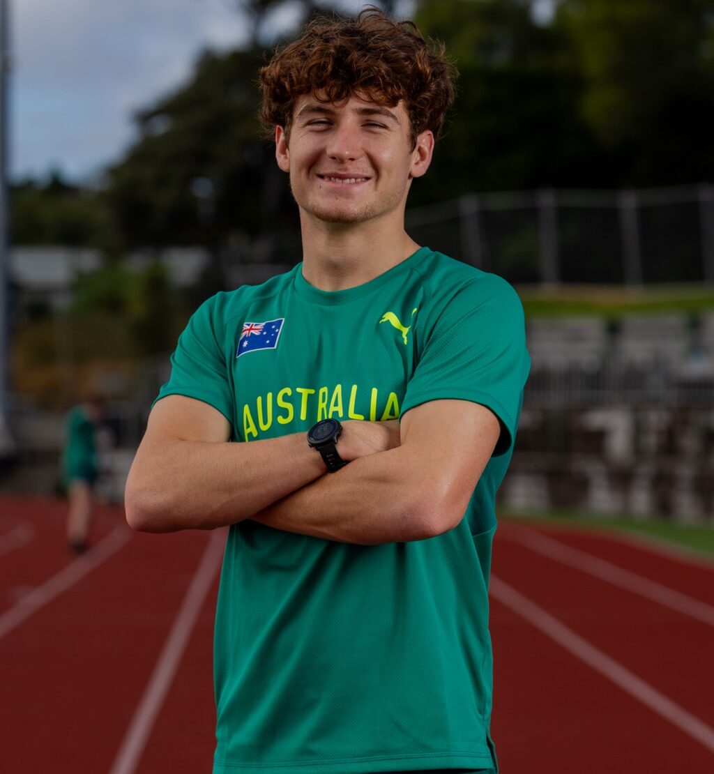 Young man with curly hair in a teal Australia shirt, arms crossed, smiling on a running track.