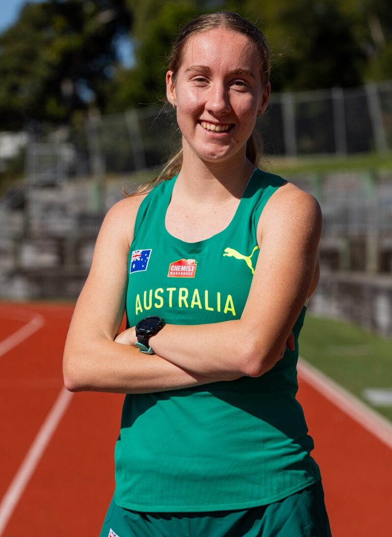 Australian female sprinter on a red track, arms crossed and smiling in a green Australia jersey on a sunny day.