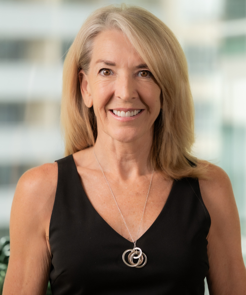 Smiling middle-aged woman with shoulder-length blonde hair wearing a black sleeveless top and a silver pendant necklace in a bright indoor setting.