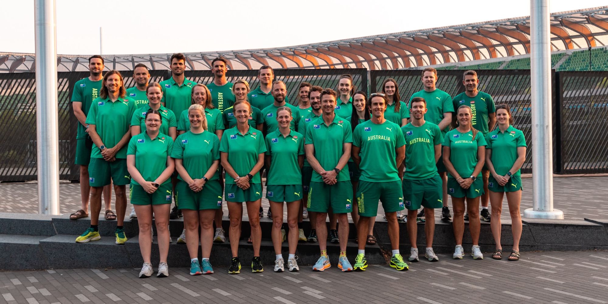 Group of Australian athletes in green uniforms posing on stadium steps outdoors.